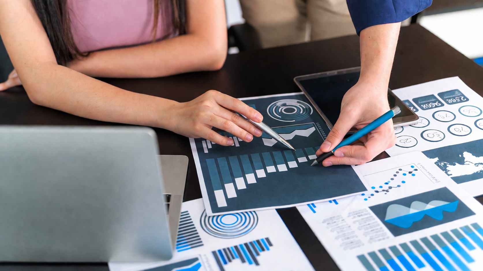 Primer plano de dos personas en una mesa de madera señalando gráficos financieros y reportes de Business Analytics durante una reunión estratégica