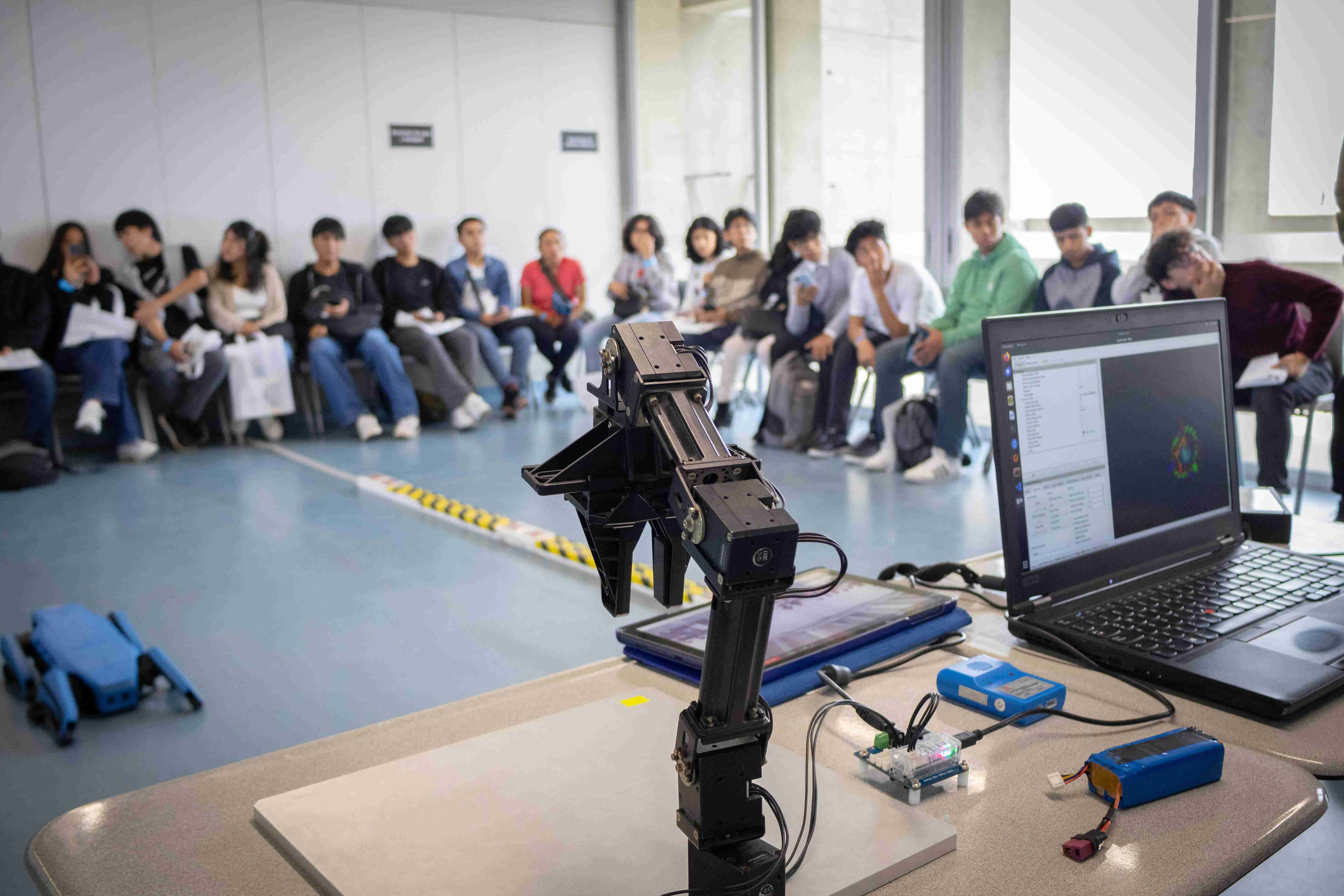 Un brazo robótico en primer plano conectado a una laptop en un laboratorio, con un grupo de estudiantes sentados al fondo observando una demostración práctica de carreras STEM